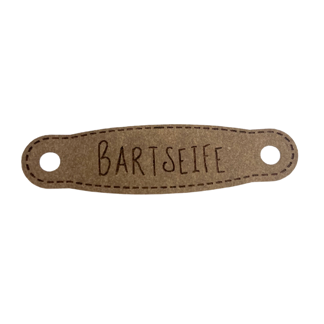 Hier ist das Etikett mit der Schrift "Bartseife" zu sehen. Es ist eine ovale Form mit zwei Löcher an der linken und rechten Seite.