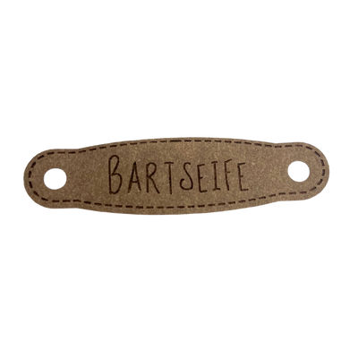 Hier ist das Etikett mit der Schrift "Bartseife" zu sehen. Es ist eine ovale Form mit zwei Löcher an der linken und rechten Seite.