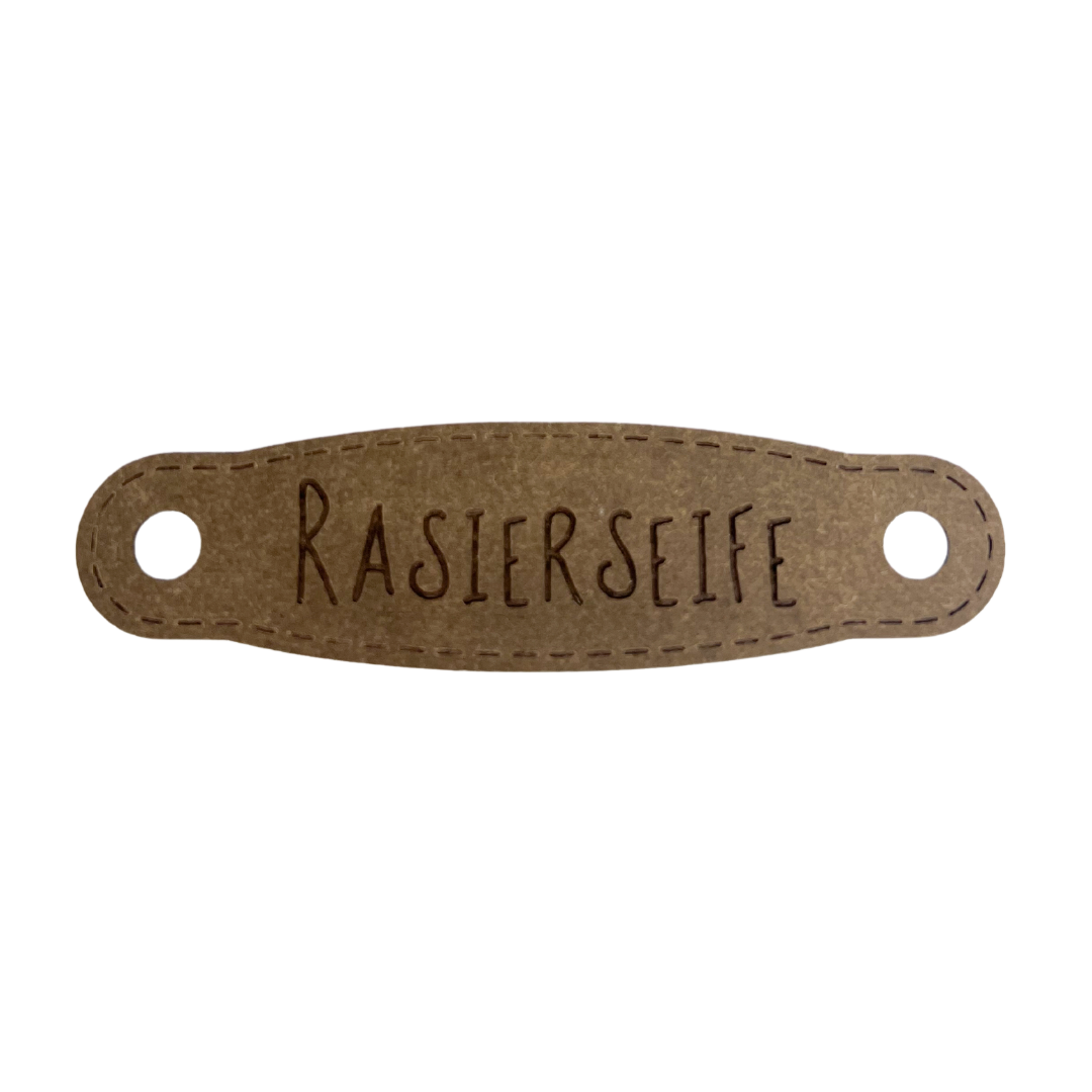 Hier ist das Etikett mit der Schrift "Rasierseife" zu sehen. Es ist eine ovale Form mit zwei Löcher an der linken und rechten Seite.