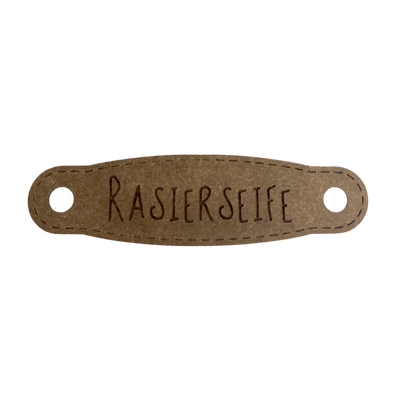 Hier ist das Etikett mit der Schrift "Rasierseife" zu sehen. Es ist eine ovale Form mit zwei Löcher an der linken und rechten Seite.
