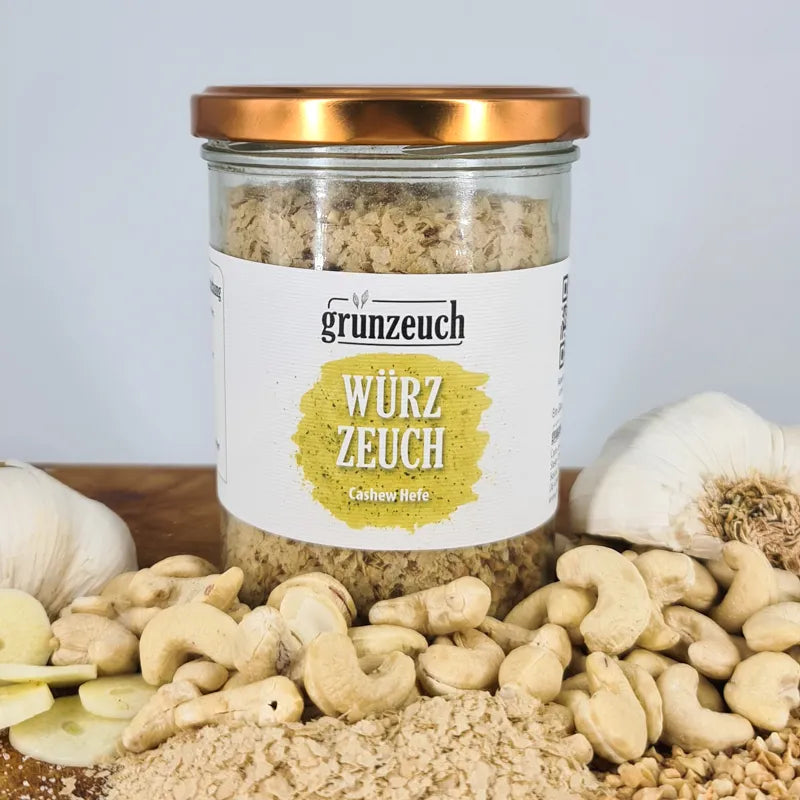 Hier kann man das Cashew Würzzeuch  im Glas sehen. Es steht auf einem Holztisch mit weißer Wand im Hintergrund. Vor dem Glas sind Cashews und Hefefloken. Rechts und Links liegen dann noch zwei Knoblauchhälften.